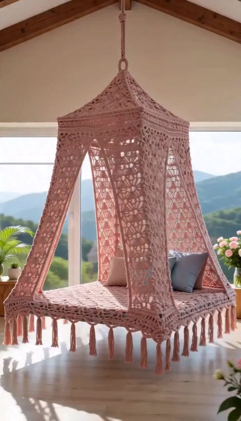 A pink hanging swing bed with tassel fringe, suspended in a rustic room with wooden ceiling beams, plants, and light wood flooring.