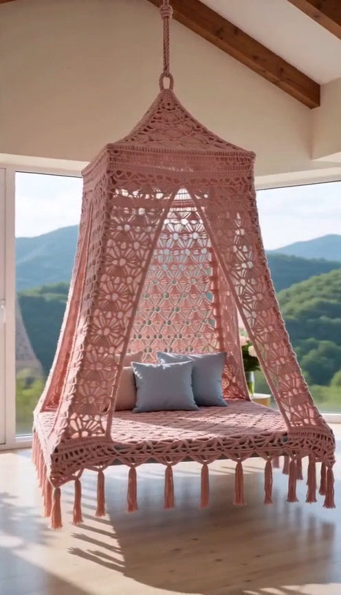 A pink hanging swing bed with tassel fringe, suspended in a rustic room with wooden ceiling beams, plants, and light wood flooring.