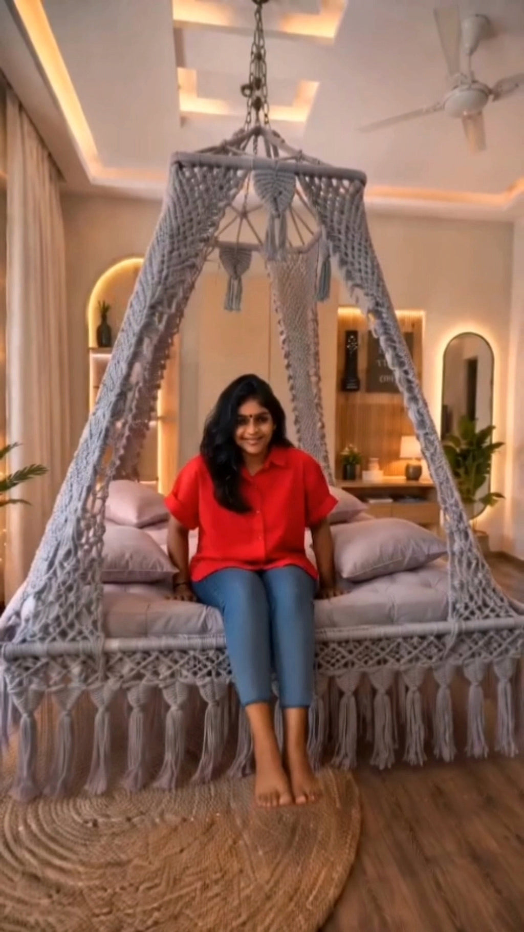 A woman sits in a large woven macrame hanging swing bed with a canopy frame, styled in a warm, modern bedroom interior.