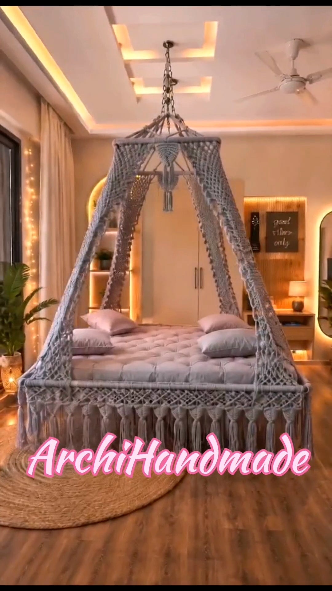 A large gray macrame canopy  swing bed frame with a tufted mattress and pillows, shown in a decorated bedroom setting with warm lighting and a wooden floor. 