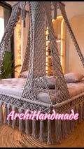 A gray macrame canopy bed net with tassels draped over a bed in a warmly lit bedroom. 