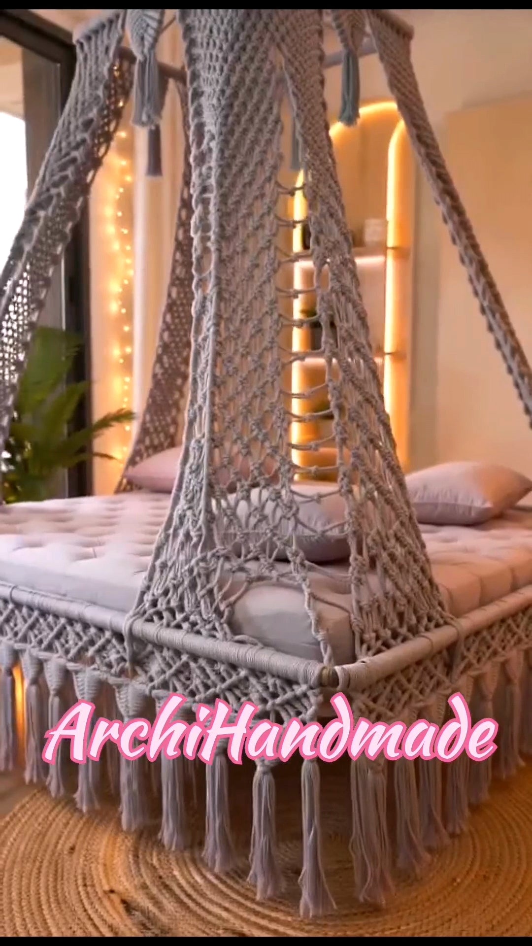A gray macrame canopy bed net with tassels draped over a bed in a warmly lit bedroom. 